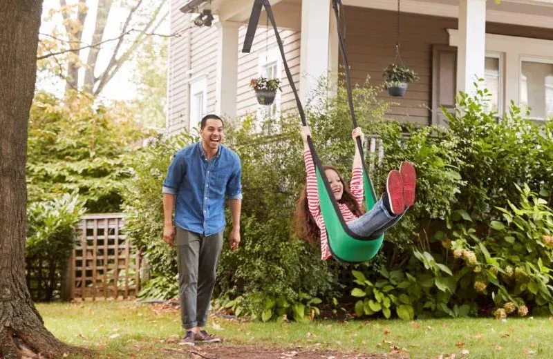 dad pushing daughter on swing in a tree in front of house